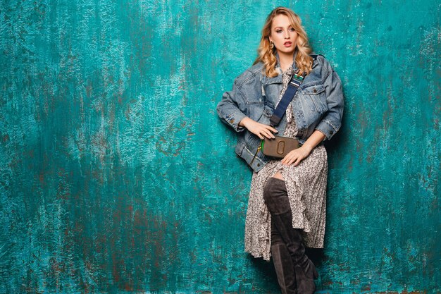 Sexy attractive stylish blonde woman in jeans and oversize jacket walking against vintage green wall in street