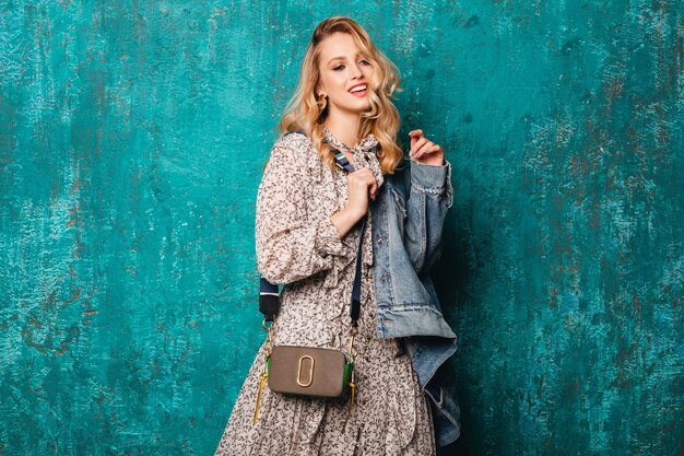 Sexy attractive stylish blonde woman in jeans and oversize jacket walking against vintage green wall in street