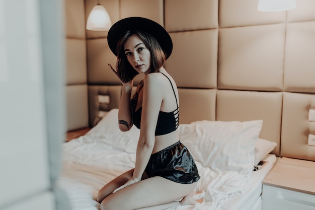 Бесплатное фото Сексуальная красивая женщина в модном черном нижнем белье и широкой шляпе сидит утром на кровати