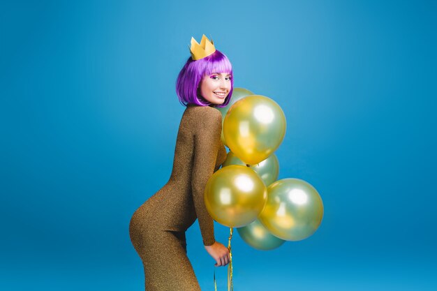 Sexy beautiful young woman in fashionable luxury dress having fun with golden balloons . Cut purple hair, crown, celebrating new year party, birthday, smiling, happiness.