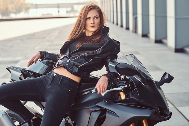 A sexy biker girl wearing a black leather jacket posing on her superbike outside a building.