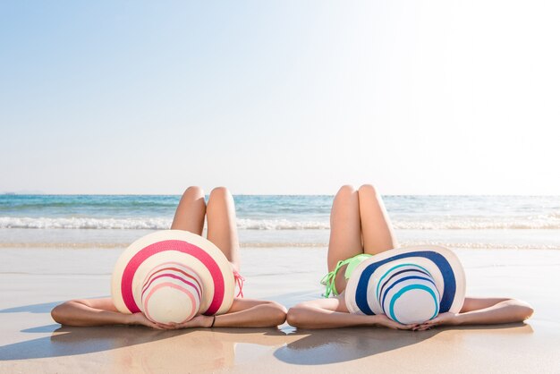 Sexy bikini body asian women enjoy the sea by laying down on sand of beach wearing millinery hat, and both legs up in the air. Happy island lifestyle. White sand and crystal sea of tropical beach.