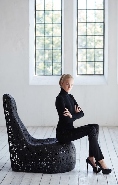 무료 사진 검은 의상을 입은 섹시한 금발 여성이 빈 방에 있는 둥근 검은 의자에 앉아 있습니다.