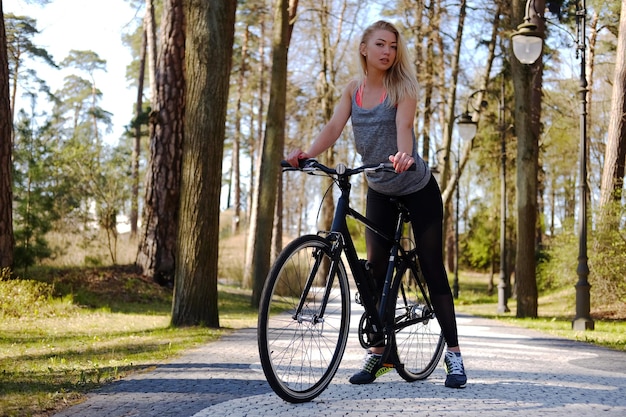 Sexy blond female posing near bicycle in a summer park.