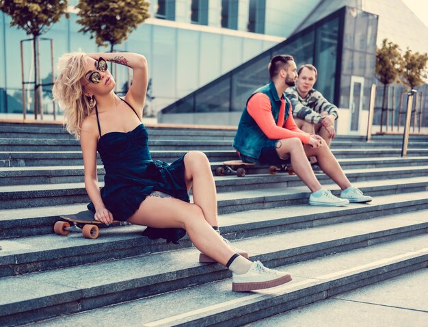 Sexy blond female with tattoo on her legs in a black shirt dress and two casual guys posing on longboard in downtown