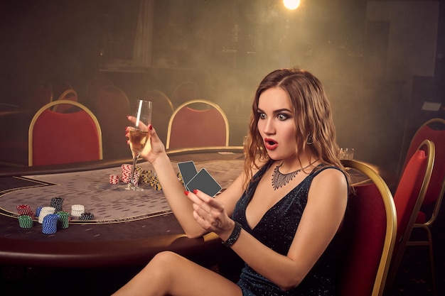 Free photo sexy blond lady in a short black dress, with playing cards and a glass of champagne in her hands is looking surprised while posing sitting sideways on a chair against a poker table in luxury casino. p