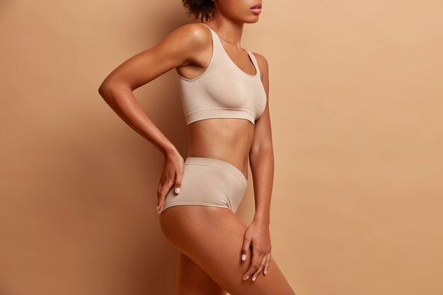 Sexy body of mixed race woman standing in profile keeps hand on buttocks dressed in underwear has perfect shining skin slim figure