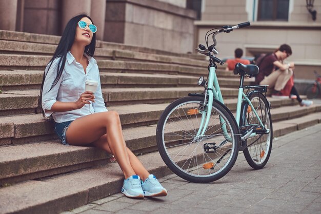 Сексуальная брюнетка в блузке и джинсовых шортах в солнцезащитных очках, расслабляющаяся после езды на велосипеде, наслаждающаяся кофе на ступеньках в городе.