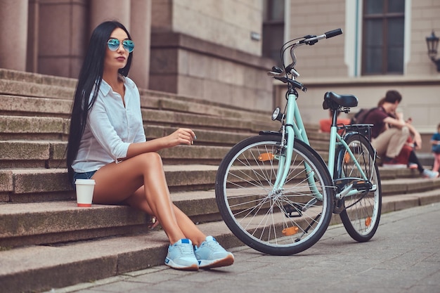 선글라스에 블라우스와 데님 반바지를 입은 섹시한 갈색 머리 여성, 자전거를 탄 후 휴식을 취하고 도시의 계단에서 커피 한 잔과 함께 앉아 있습니다.