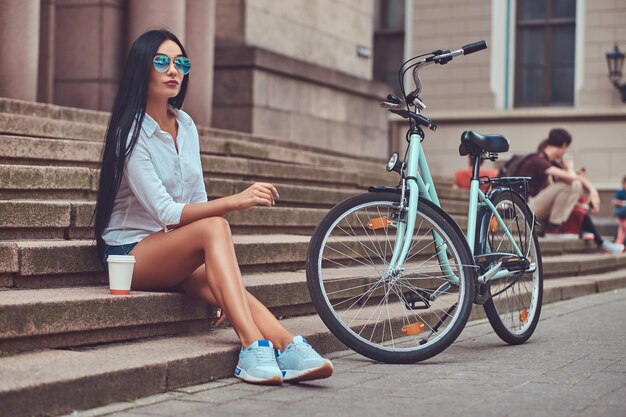 A sexy brunette female wearing blouse and   denim shorts in sunglasses, relaxing after riding on a bicycle, sitting with a cup of coffee on steps in a city.