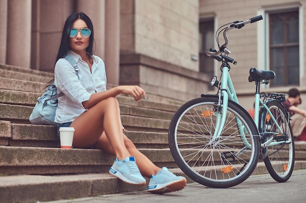 A sexy brunette female wearing blouse and   denim shorts in sunglasses, relaxing after riding on a bicycle, sitting with a cup of coffee on steps in a city.