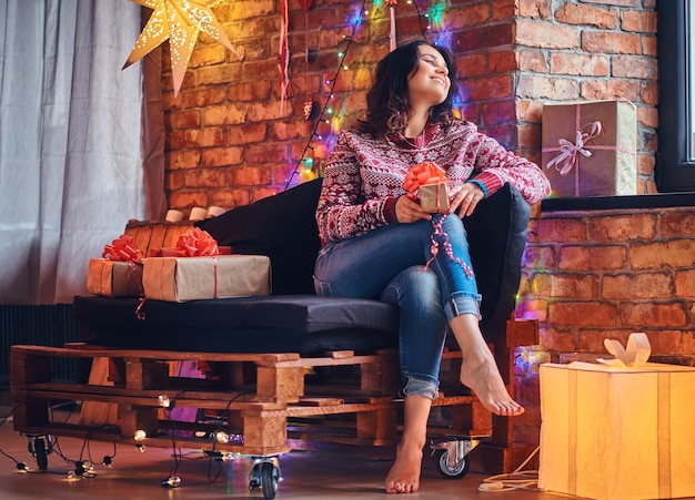 Sexy brunette female with naked foot dressed in a jeans and a red sweater posing on a sofa in a room with Christmas decoration.