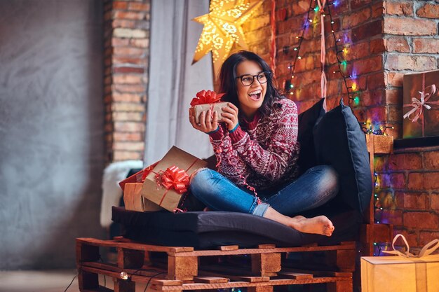 クリスマスの装飾が施された部屋のソファでポーズをとるジーンズと赤いセーターに身を包んだ裸の足でセクシーなブルネットの女性。
