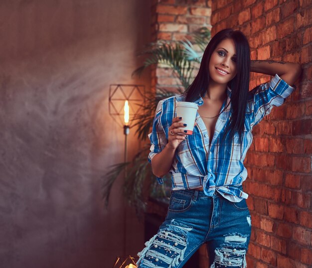 A sexy brunette in a flannel shirt and jeans holds a cup of coffee posing against a brick wall.