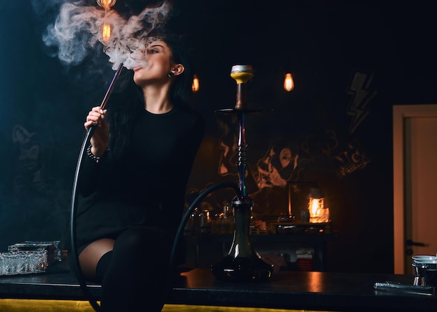 Sexy brunette girl in seductive black clothes smokes a hookah while sitting on counter in a nightclub.