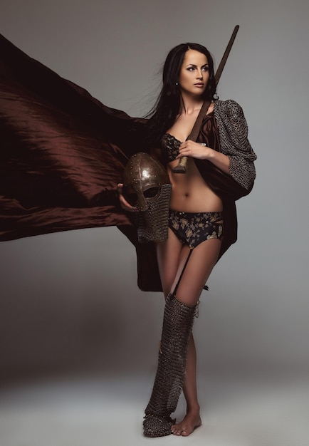ヘルメットと剣を保持している古代の鎧のセクシーなブルネットの女性。灰色の背景に分離。