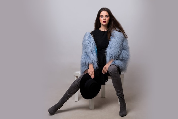 Free photo sexy brunette woman in trendy  blue fur jacket and velvet thight high boots posing  on grey wall.