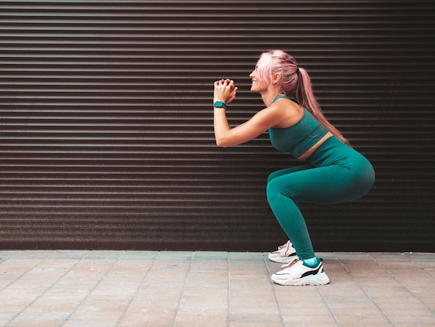 Сексуальная фитнес-женщина в зеленой спортивной одежде с розовыми волосамиОна делает приседания Молодая красивая модель с идеальным теломЖенщина на улице возле стены с рольставнямиВеселый и счастливый на улице