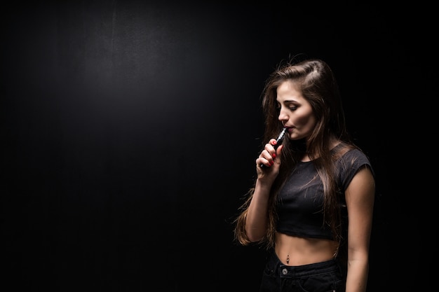 Сексуальная девушка в черном платье курит электронную сигарету на темной стене