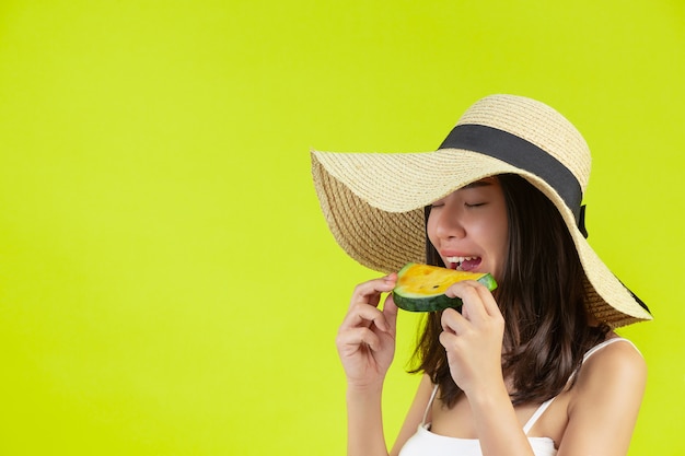 무료 사진 섹시 한 여자는 노란색 벽에 모자 sumer 더운 날씨에 watermalon를 먹고있다.