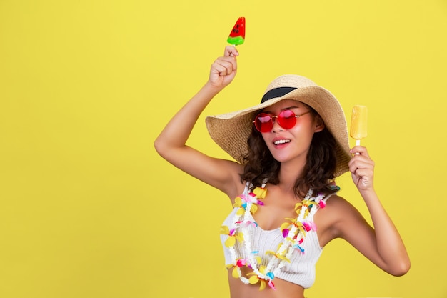 섹시한 여자는 노란색 벽에 여름 더운 날씨에 안경과 모자를 착용하는 동안 수박과 망고 아이스크림을 들고있다.