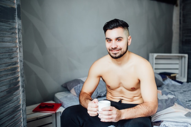 Sexy handsome young man laying shirtless on his bed, holding a coffee cup