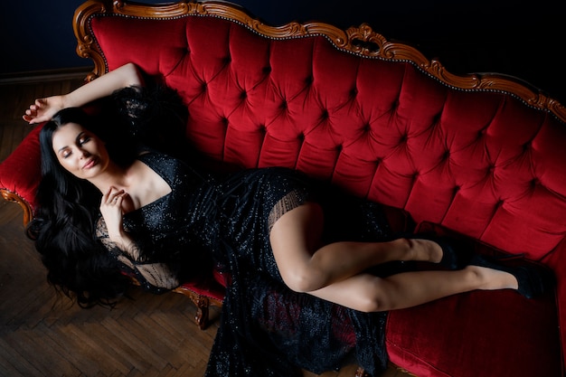 Sexy longhaired brunette caucasian girl with closed eyes is lying on the luxury red sofa dressed in black lace dress