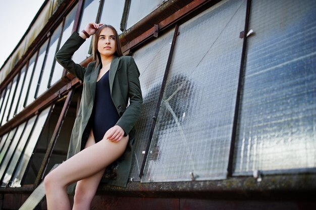 검은 란제리 복장 바디 수영복 콤비드레스와 재킷에서 긴 다리를 가진 섹시한 모델 소녀가 창문이 있는 버려진 산업 장소의 지붕에서 포즈를 취했습니다.