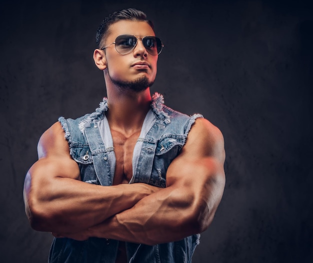 Sexy naked fashionable athletic man in a denim vest and sunglasses poses with crossed arms in a studio. Isolated on dark background.