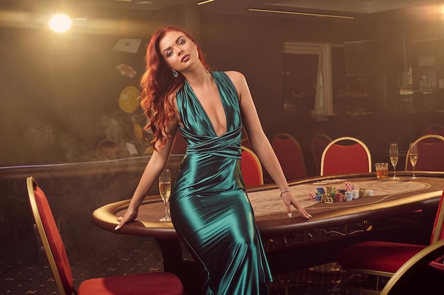 Sexy redheaded female in a long blue satin dress is posing sitting on a poker table and looking at the camera in luxury casino. Passion, cards, chips, alcohol, win, gambling - it is as female entertai