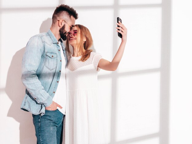 Sexy smiling beautiful woman and her handsome boyfriend Happy cheerful family having tender moments near white wall in studioPure cheerful models huggingEmbracing each other Taking selfie