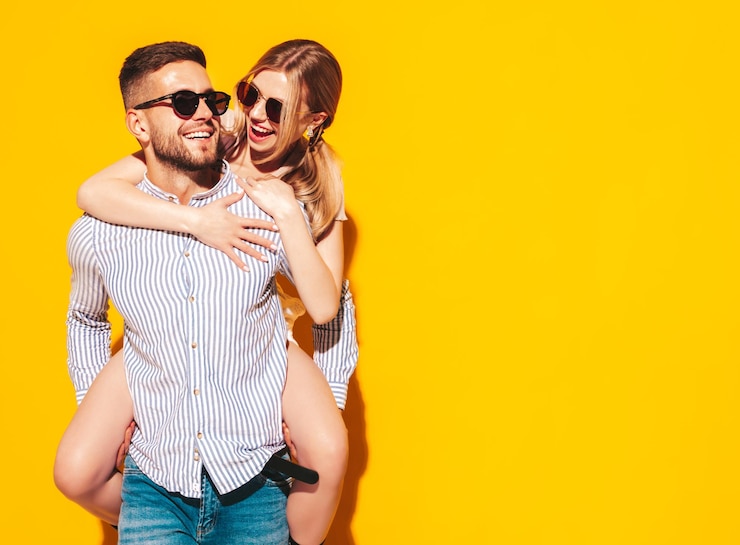 Free Photo sexy smiling beautiful woman and her handsome boyfriend happy cheerful family having tender moments near yellow wall in studiopure cheerful models gives piggyback ridingembracing in sunglasses