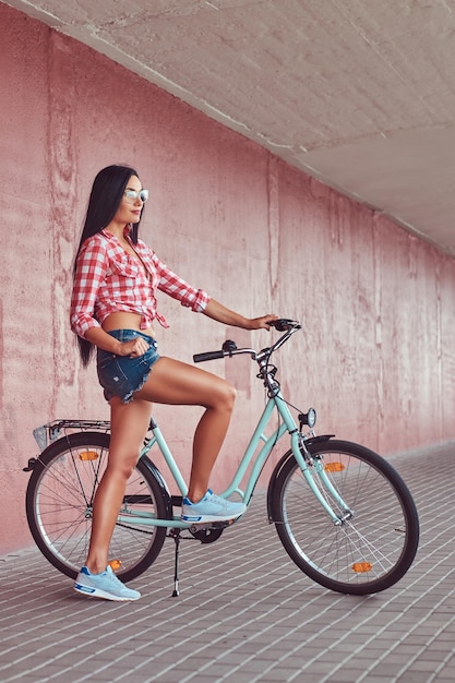 A sexy stylish brunette girl wearing a pink flannel shirt and denim shorts in glasses, posing with city bike against a pink wall.