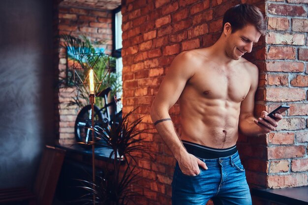 A sexy tattoed shirtless man using a mobile phone leans against the brick wall. Smiling and looking away.