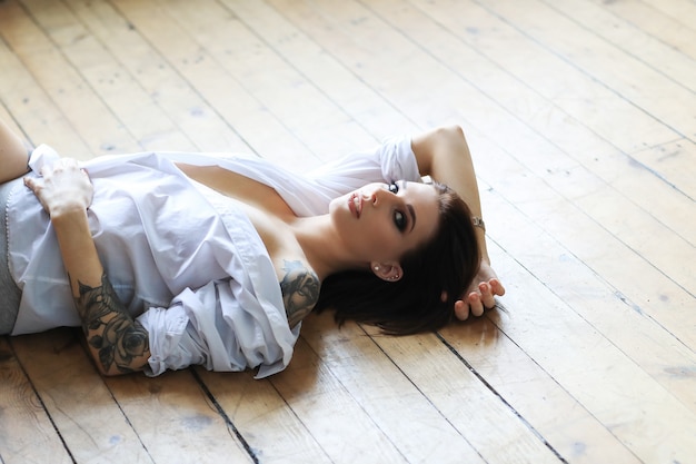 Сексуальная татуированная женщина в белой рубашке лежит на деревянном полу