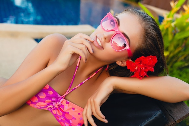 ビキニとピンクのサングラス、熱帯の花、カラフルな夏のファッションスタイルを身に着けているプールで横たわっている夏休みのセクシーな女性
