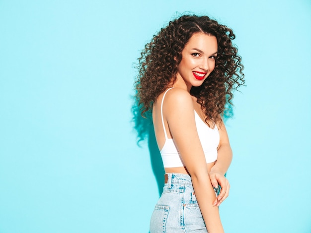 Sexy young beautiful smiling female in trendy summer hipster clothes carefree woman posing near blue wall in studio Positive brunette model with afro curls hairstyle Cheerful and happy