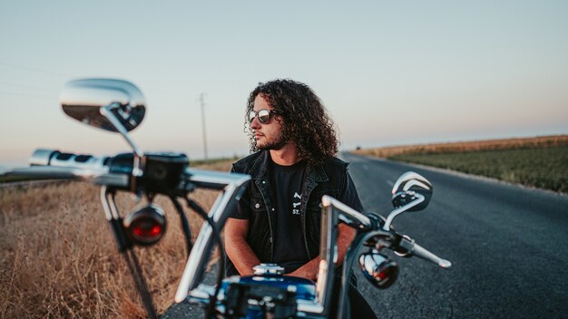 Shallow focus of a cool curly-haired male with a black denim jacket on his motorcycle on the road