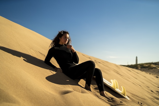 Shallow focus shot of an attractive female posing on a sandy hill with a surfboard on the side