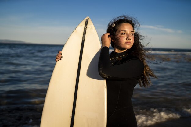 Shallow focus shot of an attractive female posing at the seashore in Spain while holding a surfboard