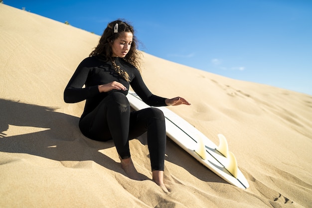 側面にサーフボードを置いて砂丘に座っている魅力的な女性の浅いフォーカスショット