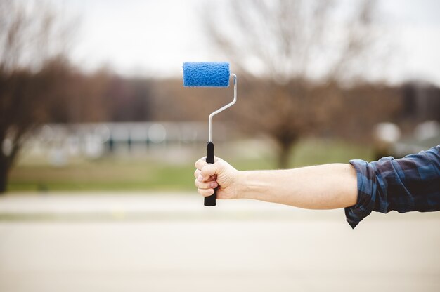 Shallow focus shot of a hand holding a blue pint brush, with a park