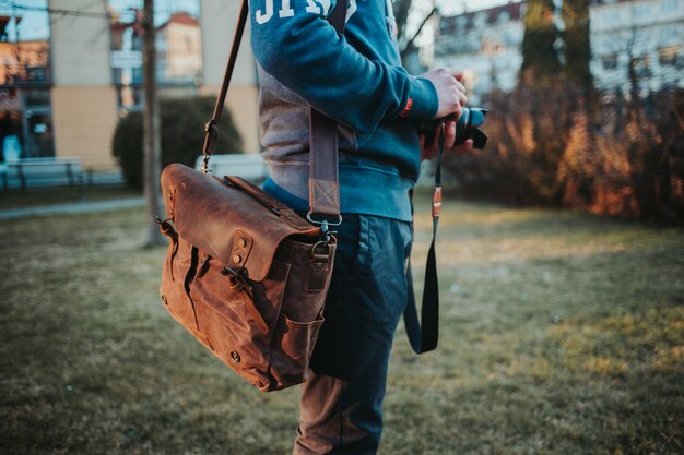 Неглубокий снимок человека с фотоаппаратом и коричневой кожаной сумкой