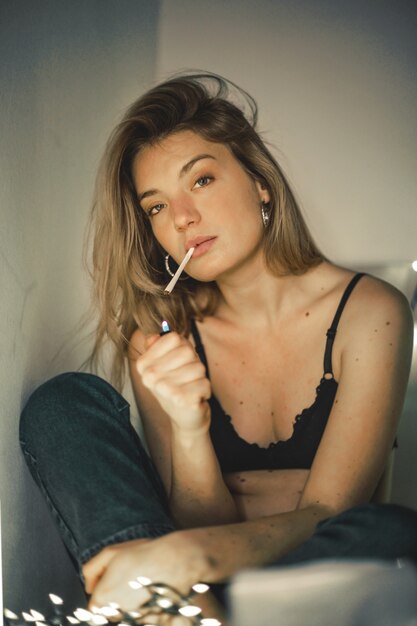 Неглубокий фокус женщины, закуривающей сигарету на кровати со светлыми гирляндами за спиной