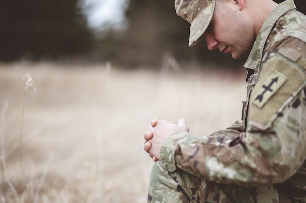Неглубокий фокус молодой солдат молится, стоя на коленях на сухой траве