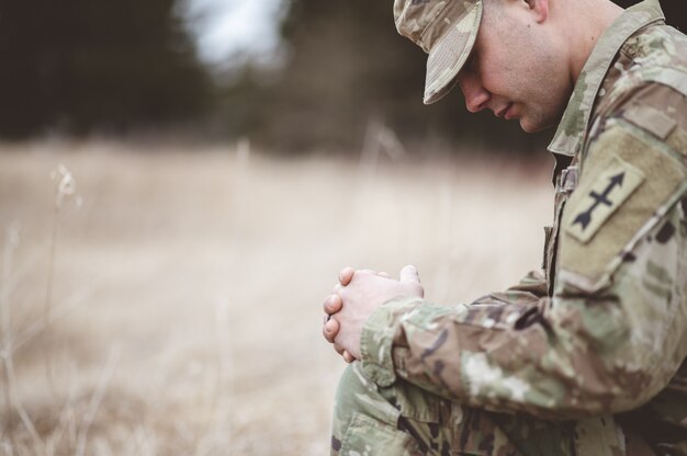 Shallow focus of a young soldier praying while kneeling on a dry grass