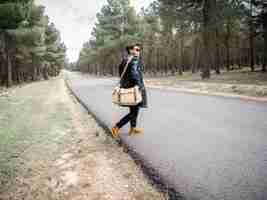 Free photo shallow focus of a young stylish man with a bag and sunglasses crossing a road in a forest