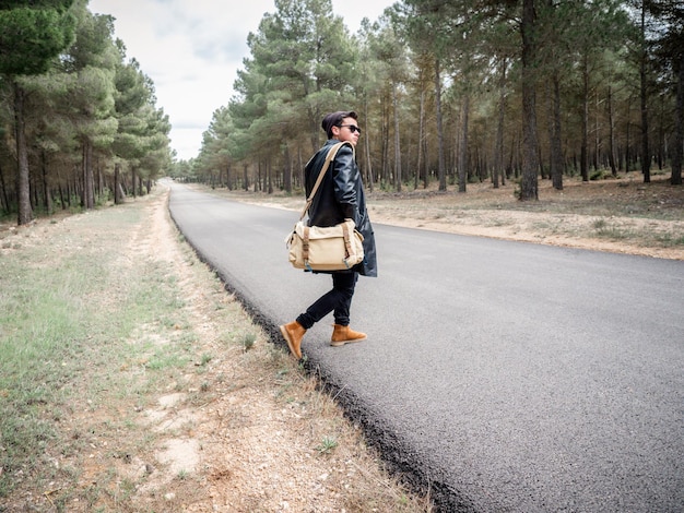 Shallow focus of a young stylish man with a bag and sunglasses crossing a road in a forest