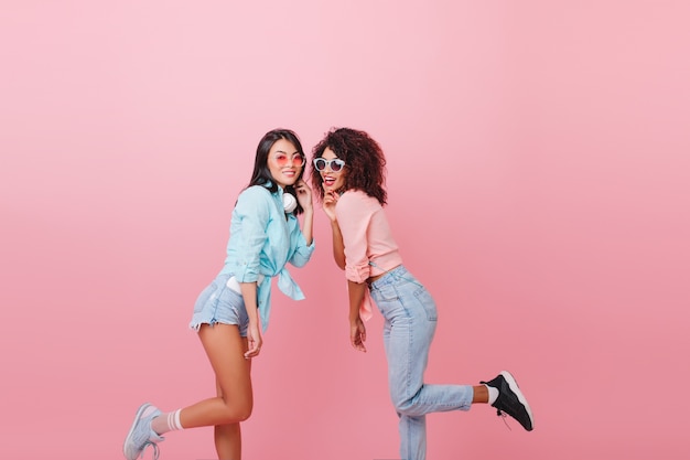 Shapely african woman in blue jeans standing on one leg, leaning to european female model. Indoor joint portrait of stunning brunette girls in casual outfit fooling around.