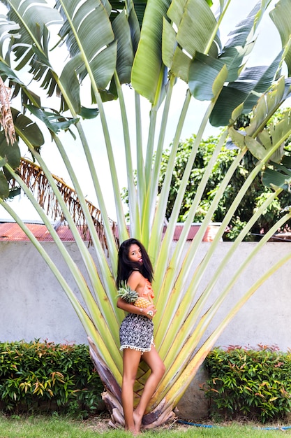 Shapely asian girl with shiny skin posing at exotic resort after sunbathing. Hispanic brunette woman in trendy bikini standing near palm tree and looking with interest.